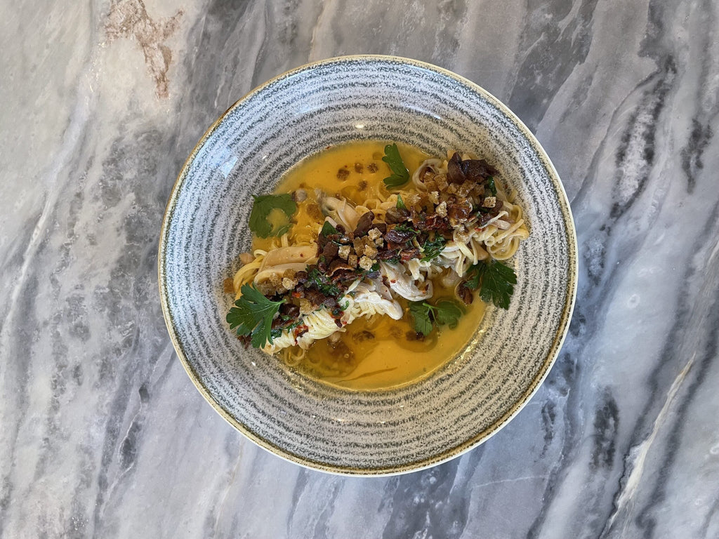 Top down view of razor clam pasta in golden broth topped with crispy lamb bacon, parsley, and breadcrumbs