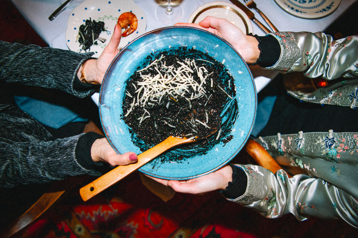 Two people passing a blue bowl of squid ink risotto topped with grated cheese and a wooden spoon at a dinner table