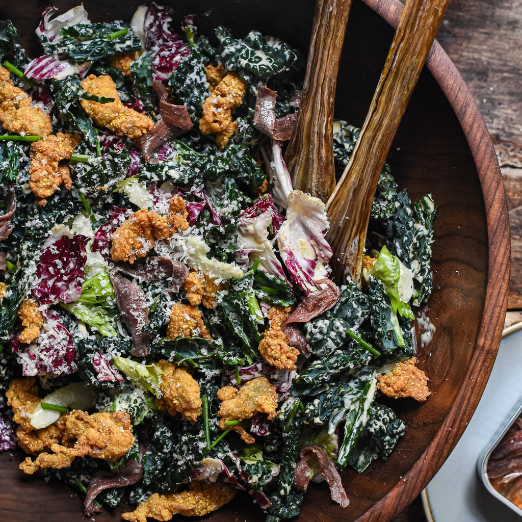 Kale salad tossed in creamy dressing, topped with fried oyster croutons, radicchio, anchovy fillets, and grated Parmesan in a wooden serving bowl