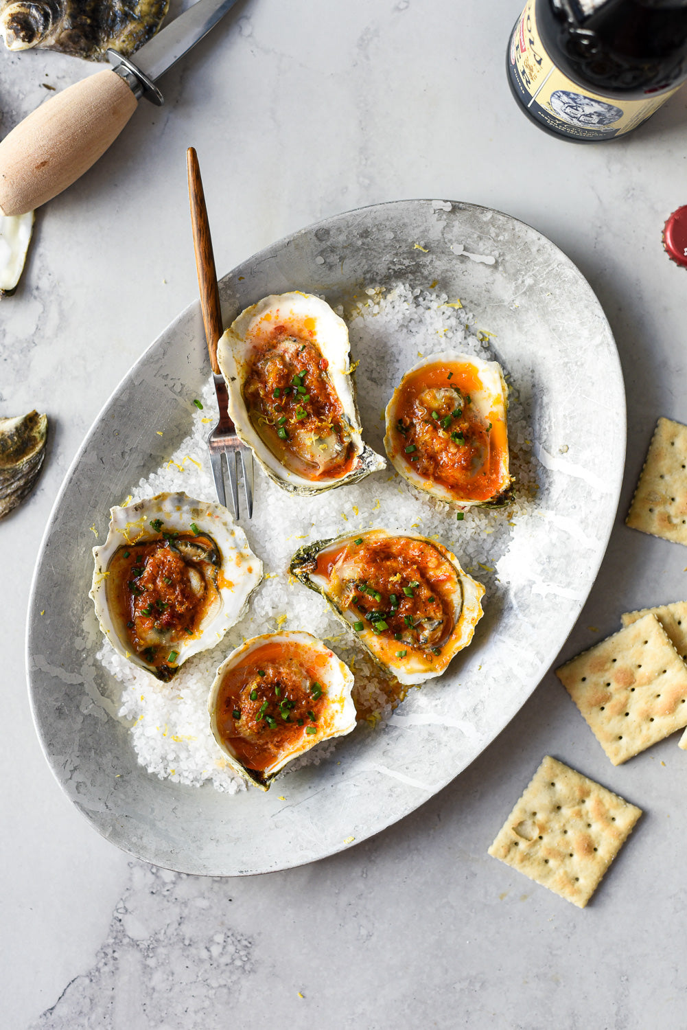 Overhead view of roasted oysters with Calabrian chile butter showcasing golden butter topping and chopped chives on coarse salt in a metal platter with crackers