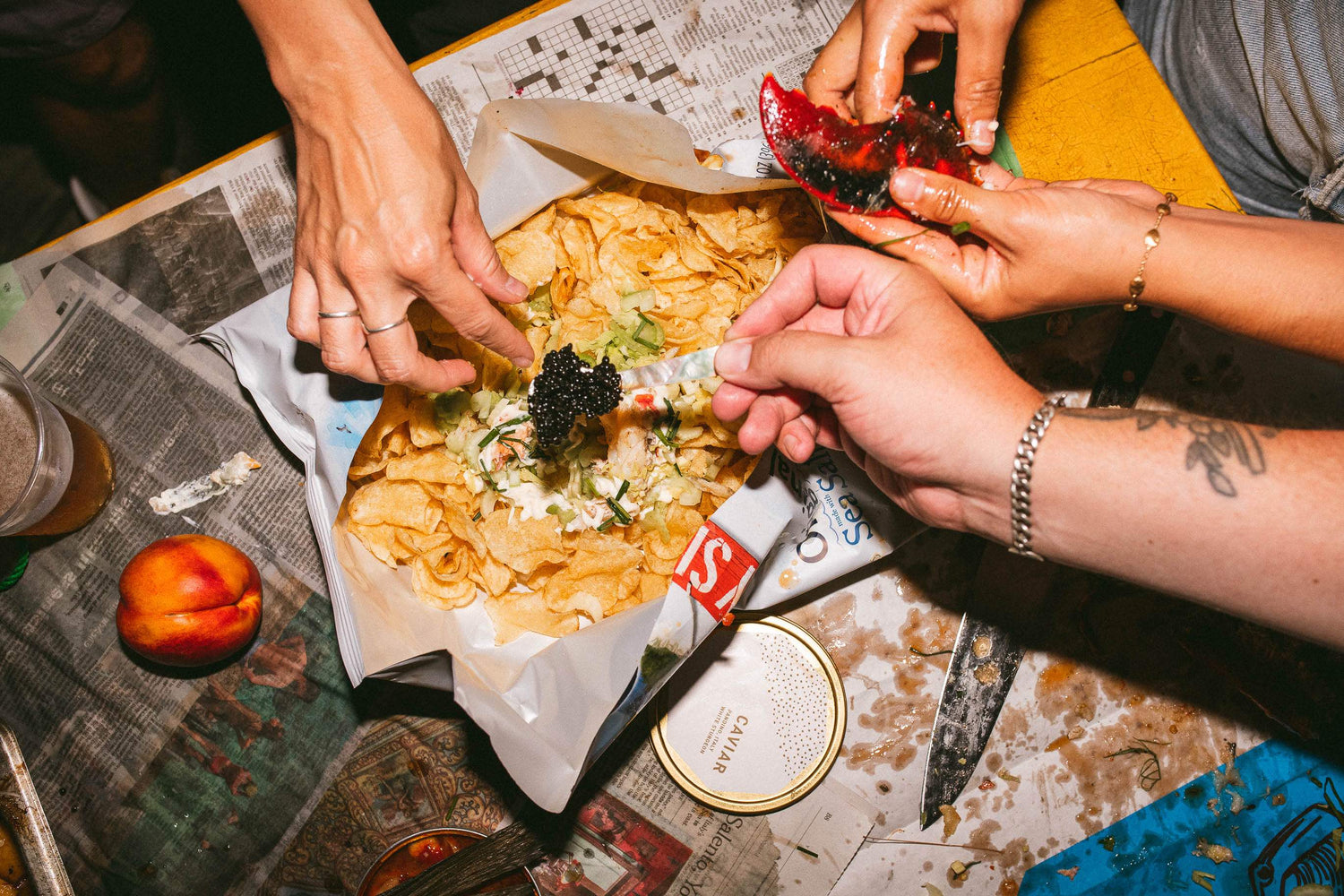 Hands assembling a lobster roll with potato chips while spooning black caviar on a newspaper-covered table