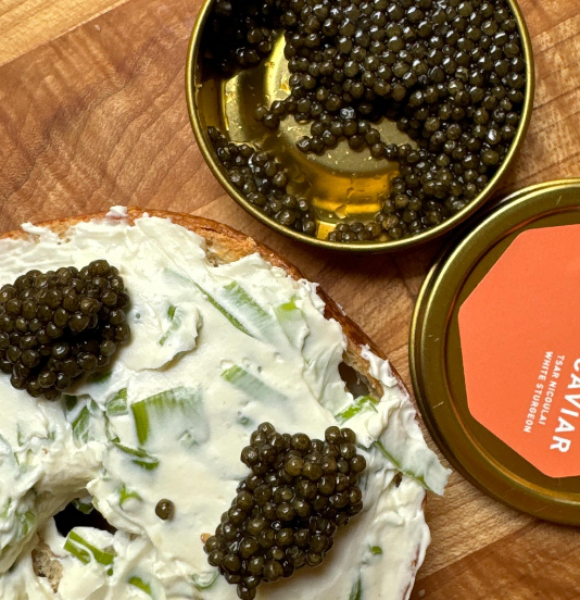 Half bagel spread with scallion cream cheese topped with three dollops of caviar beside an open caviar tin on a wooden cutting board