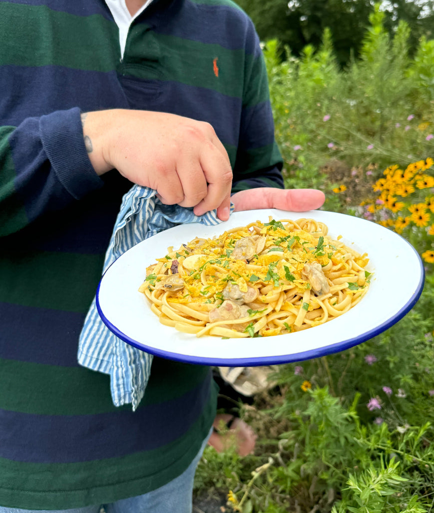 Person holding white enamel plate of pasta with tinned littleneck clams, crushed potato-chip crumbs and parsley outdoors