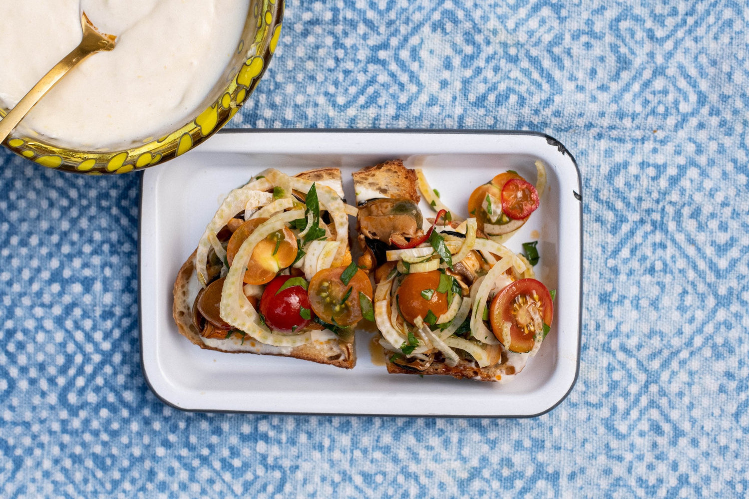 Two mussel bruschetta with cherry tomatoes and fennel on a white dish over blue cloth, lemon aioli at top left