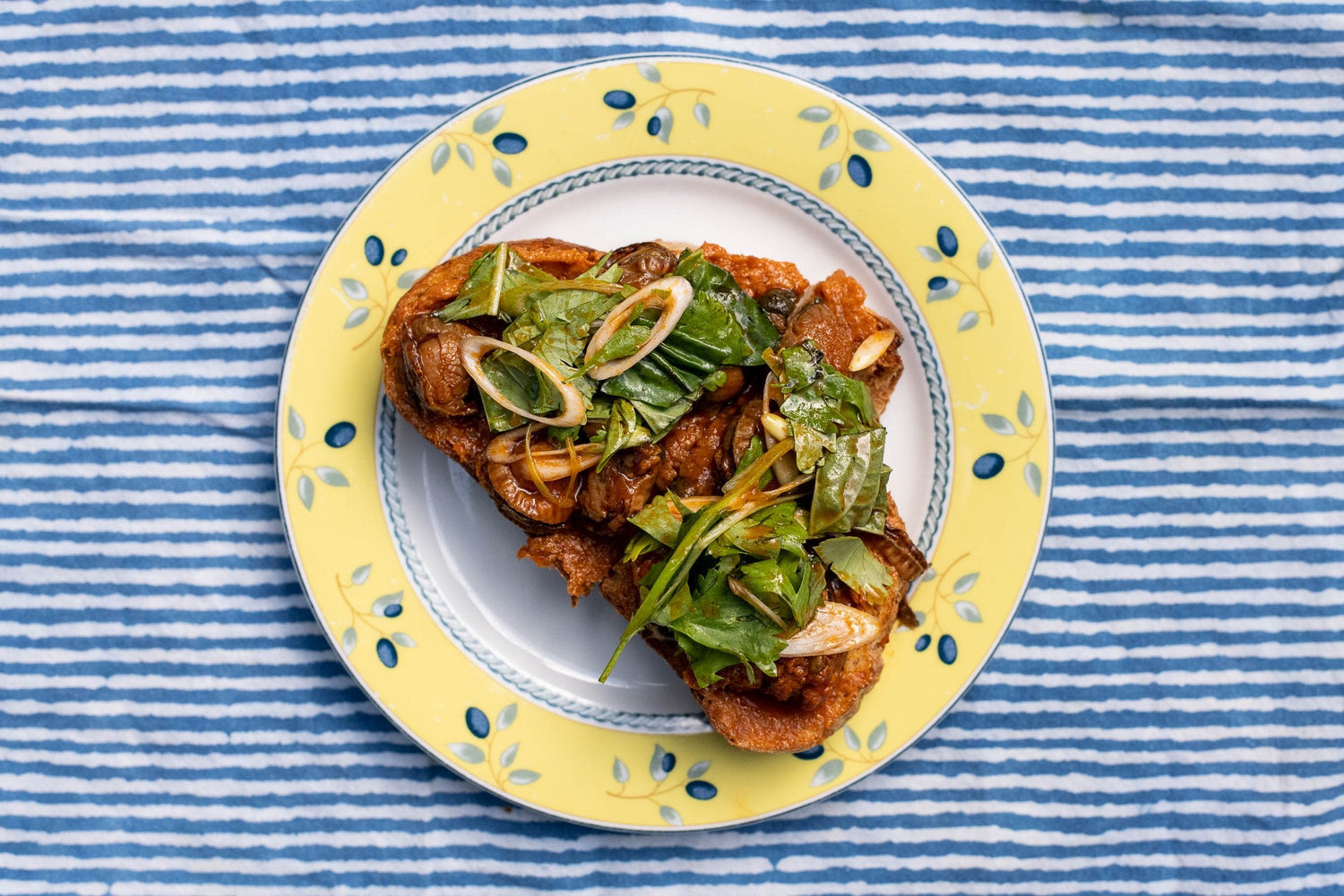 Overhead of Galician tinned scallop toast on yellow plate showing browned scallops, crisp toast and herb salad on blue-striped cloth
