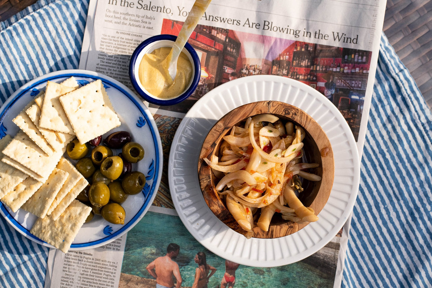 Top-down view of marinated razor clams in a wooden bowl with sliced onions, served with crackers, olives, and mustard