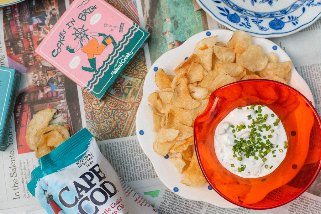 Bowl of creamy clam dip topped with chopped chives beside a plate of potato chips and tins of cockles on newspaper
