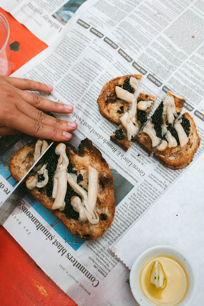 Tinned razor clams and caviar on grilled garlic toast on newspaper with a hand and a dish of olive oil and lemon