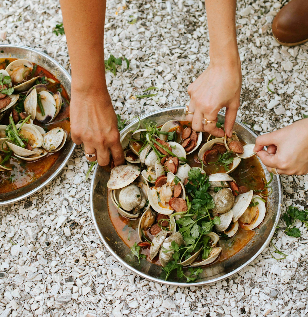 Hands serving bowls of steamed clams and sliced sausage in a spiced broth on a shell-covered driveway