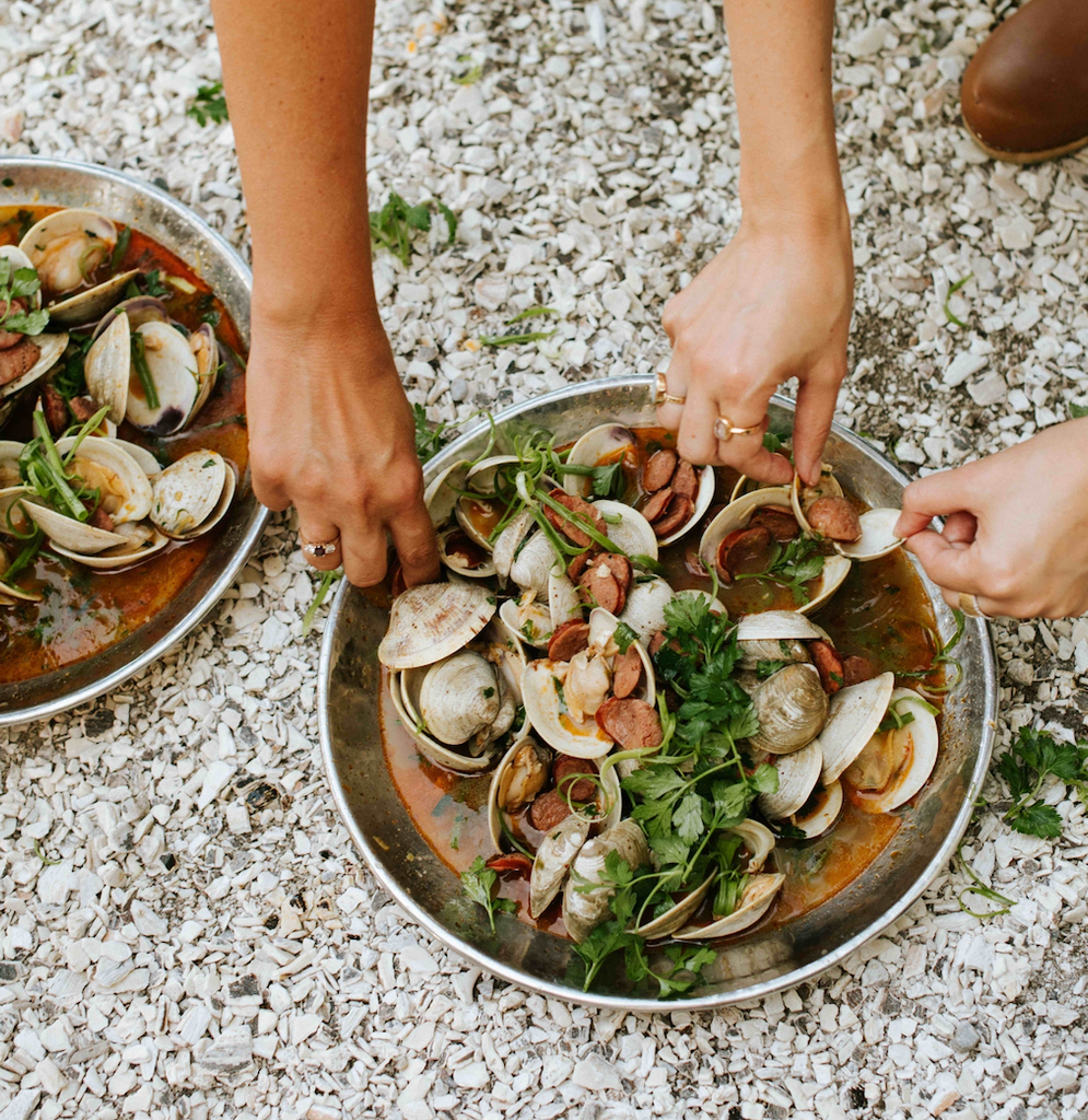 Hands serving bowls of steamed clams and sliced sausage in a spiced broth on a shell-covered driveway