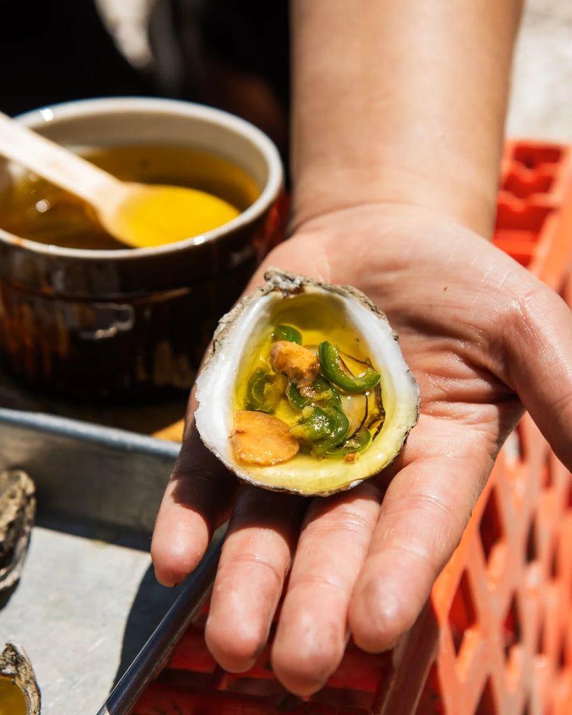 Hand holding a grilled oyster topped with olive oil, sliced green chilies, and garlic over a metal tray