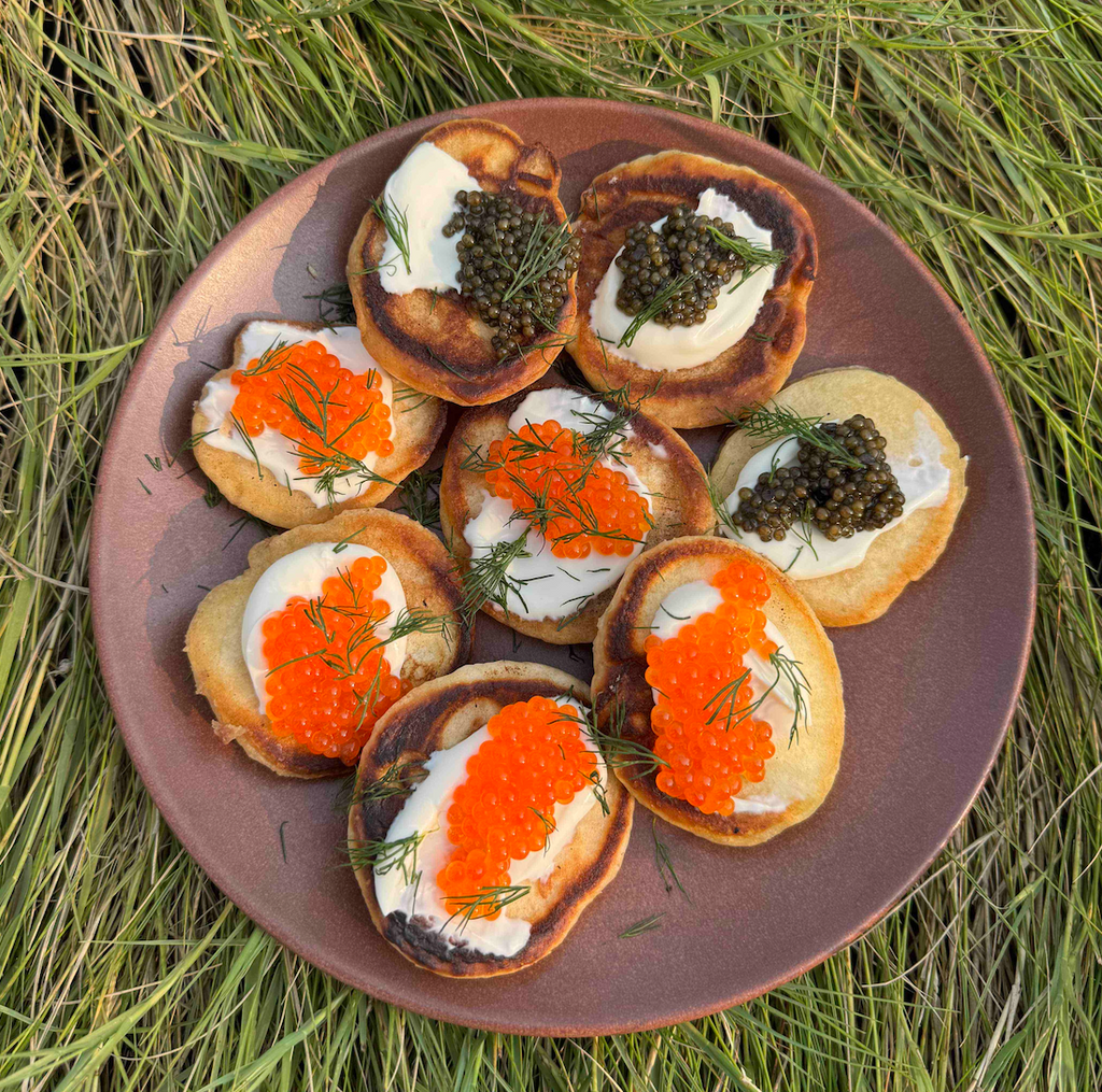 Top-down view of brown butter blinis topped with orange trout roe, black caviar and creme fraiche on a brown plate on grass