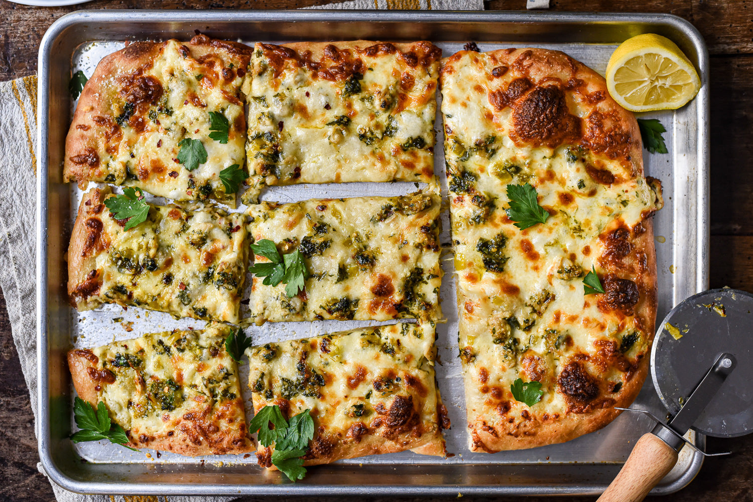 Tinned white clam pizza sliced into rectangular pieces on a baking sheet, topped with melted cheese, chopped parsley, and a lemon half