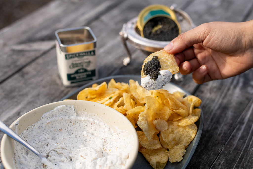 Hand holding a potato chip topped with mignonette dip and black caviar above a bowl of potato chips and a ramekin of dip on a weathered wooden table