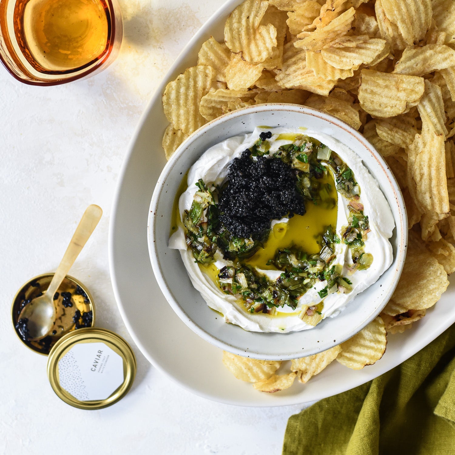 Overhead view of charred scallion sour cream dip topped with black caviar and olive oil, in a white bowl with potato chips