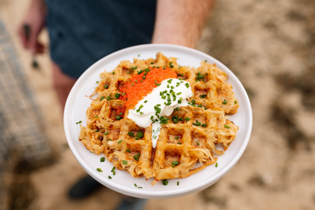 Top down view of a hash brown waffle topped with orange trout roe, a dollop of sour cream and chopped chives on a white plate held outdoors
