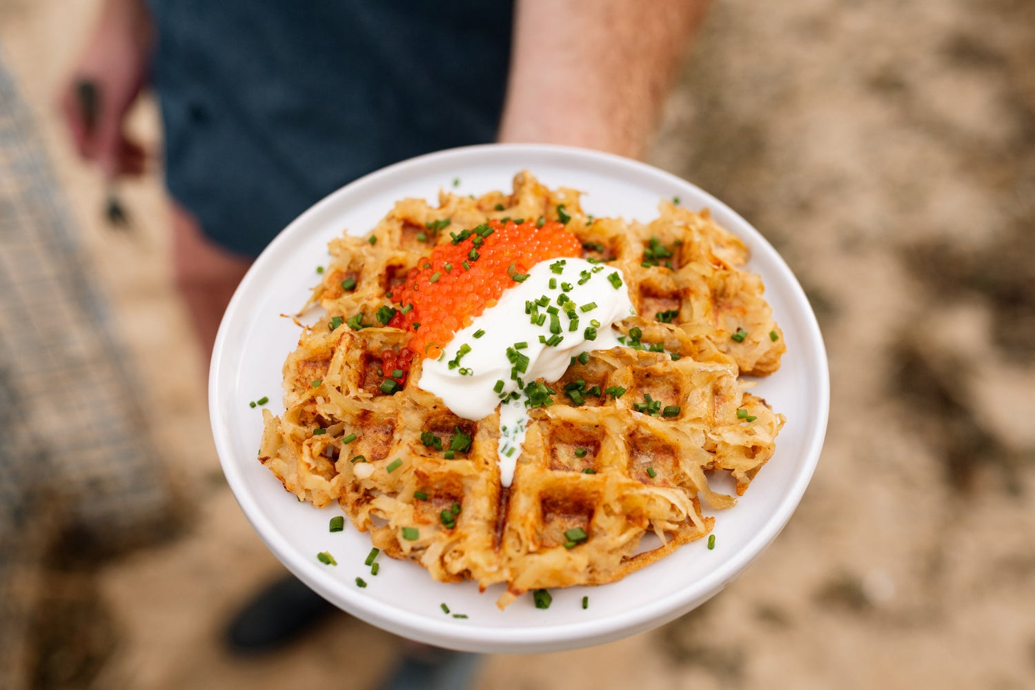 Top down view of a hash brown waffle topped with orange trout roe, a dollop of sour cream and chopped chives on a white plate held outdoors