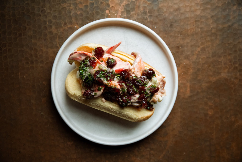 Top-down view of a lobster roll with creamy lobster salad, cranberry-herb topping and chopped chives on a toasted split roll on a white plate over a copper surface