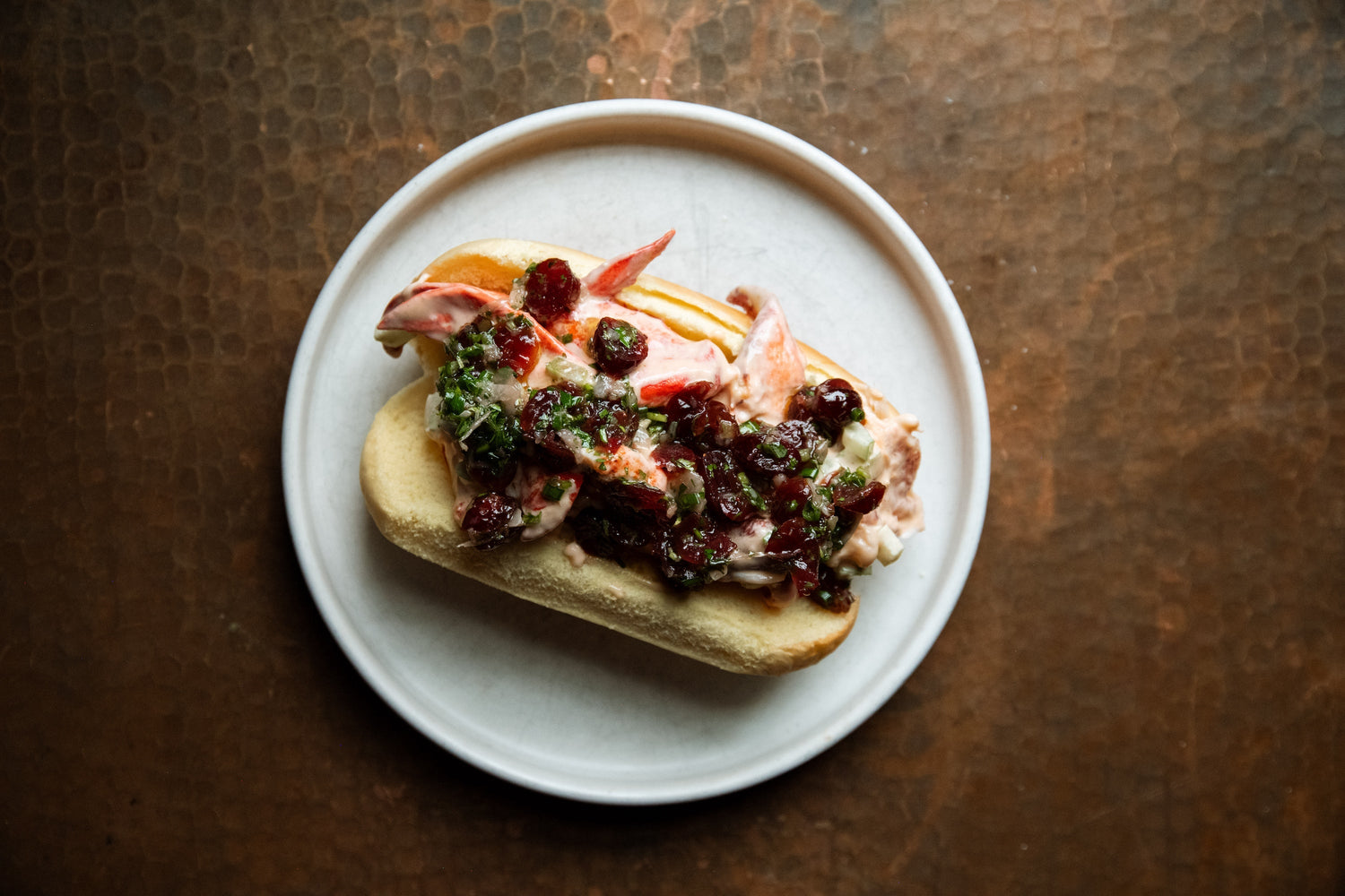 Top-down view of a lobster roll with creamy lobster salad, cranberry-herb topping and chopped chives on a toasted split roll on a white plate over a copper surface