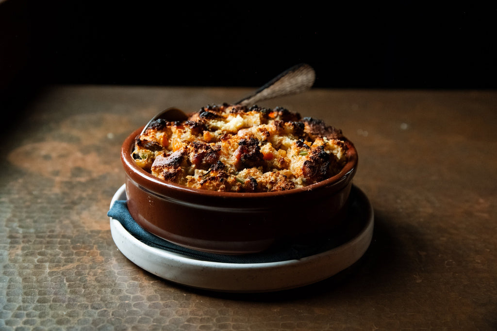 Close-up of oyster and cornbread stuffing in a brown ramekin with golden crispy topping on a dark table