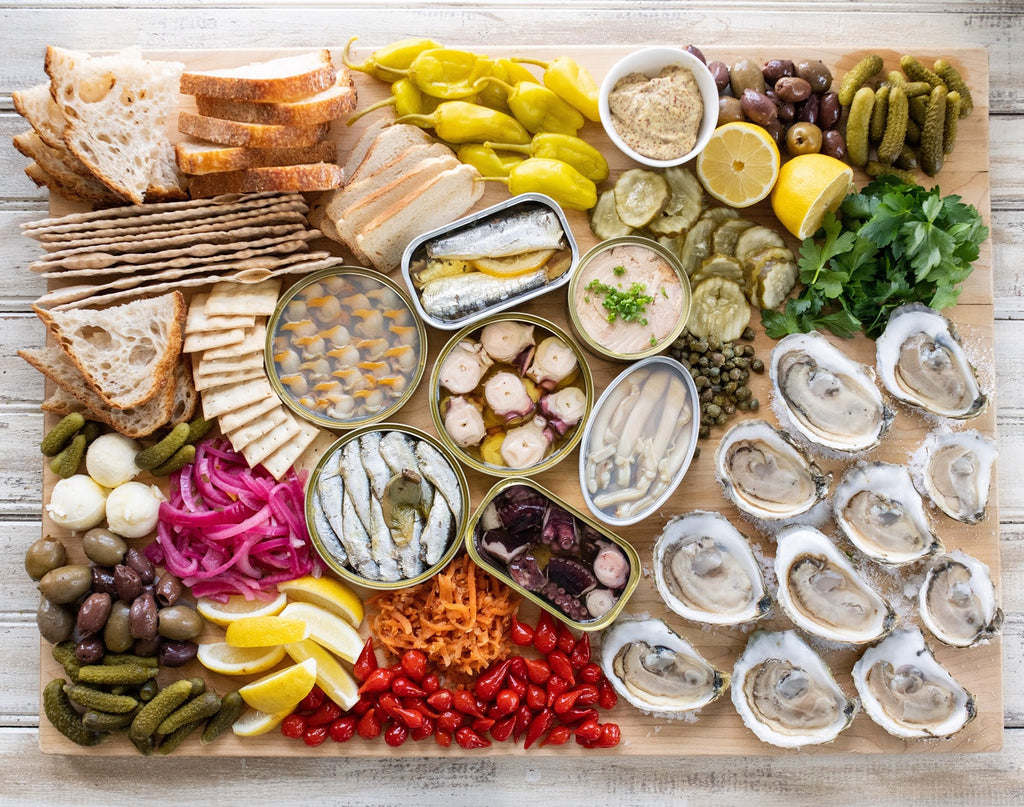 Tinned fish and oysters on a wooden board with bread, crackers, pickles, olives, lemon wedges, and herbs