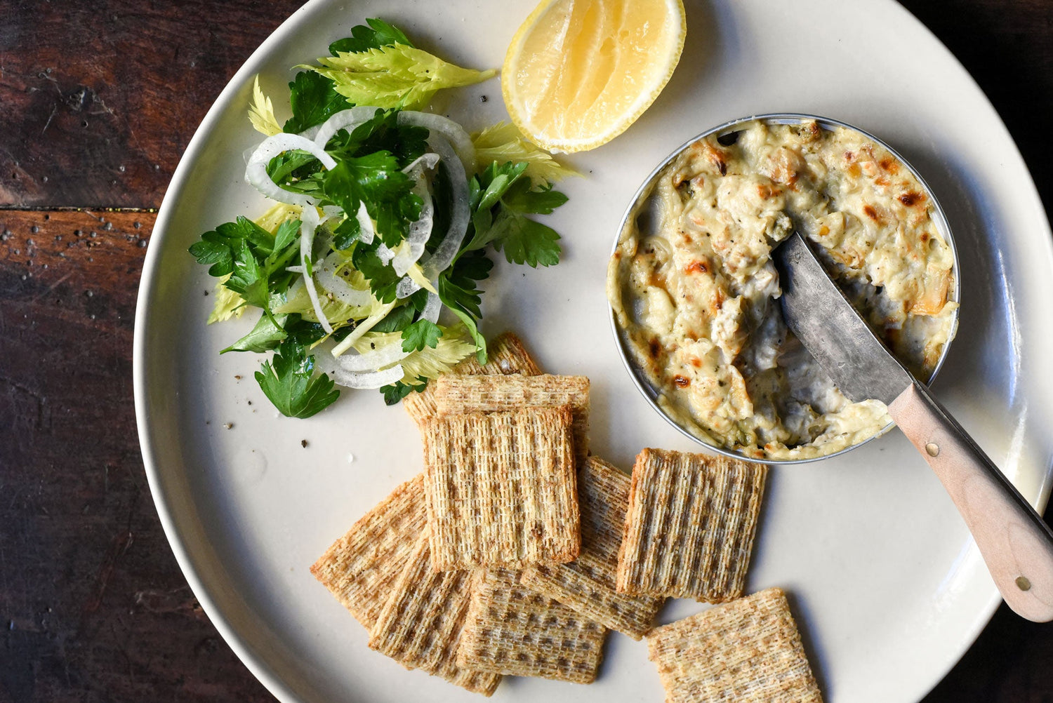 Baked cockle dip served in a small tin with a spreading knife, whole-grain crackers, and a celery and parsley salad on a white plate