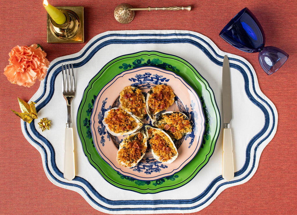 Six baked oysters with herbed breadcrumbs and bacon on stacked decorative plates, fork and knife on a placemat