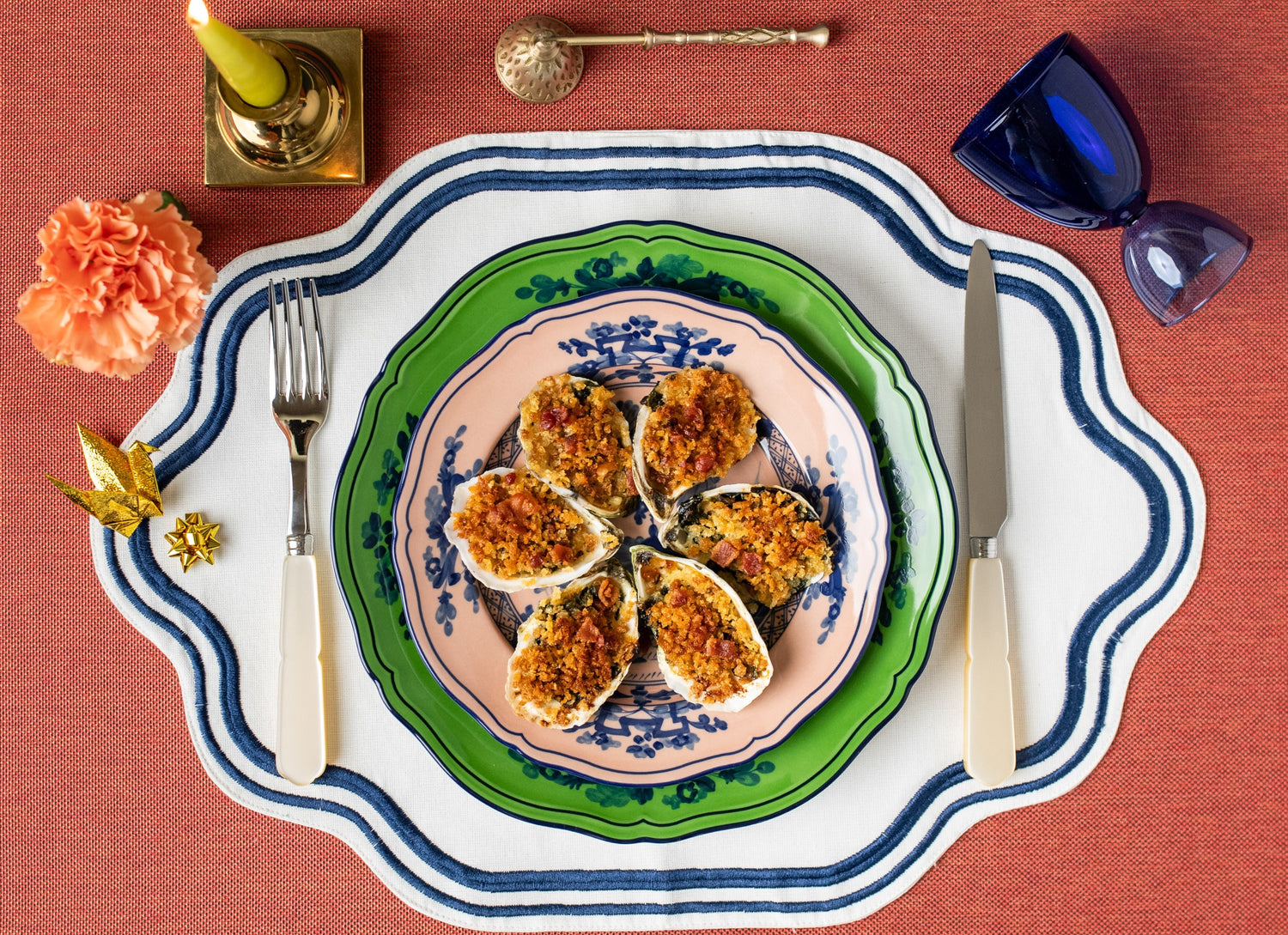 Six baked oysters with herbed breadcrumbs and bacon on stacked decorative plates, fork and knife on a placemat