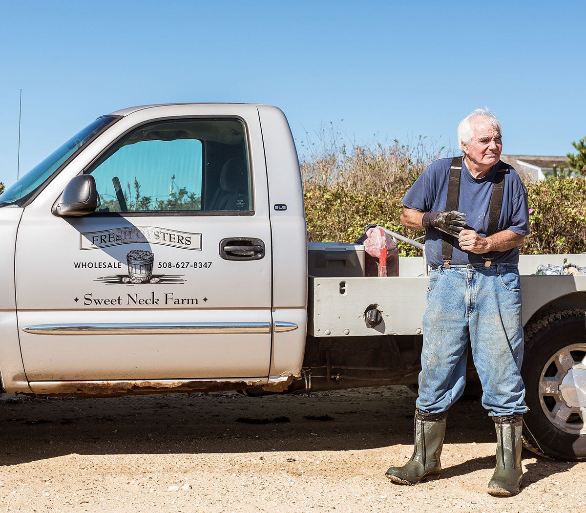 Oyster farmer in rubber boots standing beside a pickup truck labeled Sweet Neck Farm on Martha’s Vineyard shoreline